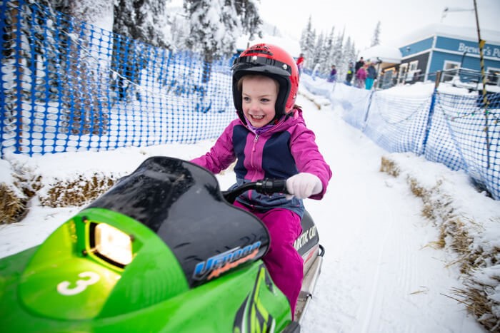 A child on a mini snowmobile going around a fenced in track