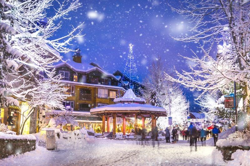 A photo of Whistler Village in early December at night. There is snow covering the walkways and the trees.