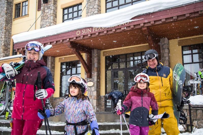 A family of four, including a mom, dad, and two girls wearing ski gear, leaving a building in the village at Sun Peaks.