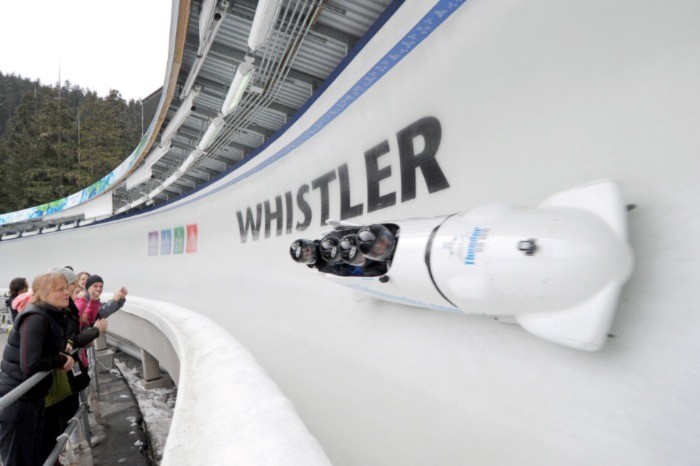 Four passengers in a bobsled going down the track with onlookers