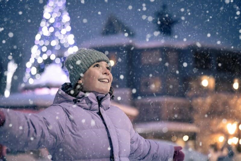 A photo of a girl wearing a grey hat and purple snow. She has her hands open and she is looking up at the snow falling in Whistler.
