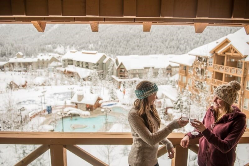 Two women wearing sweaters are clinking their glasses of wine on a balcony overlooking the hot pools at Panorama.