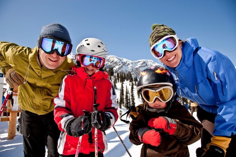 A photo of a family of four dressed in ski gear on a ski hill, looking at the camera.