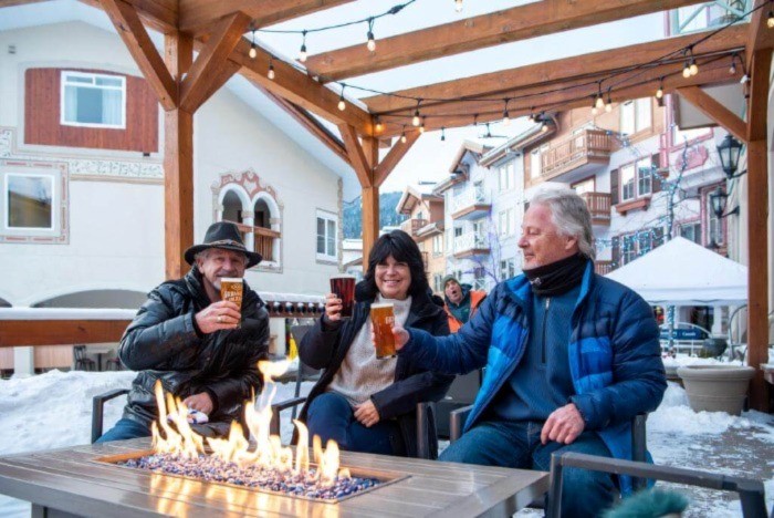 Three older adults drinking draft beer in front of a fire table on an outdoor patio in winter