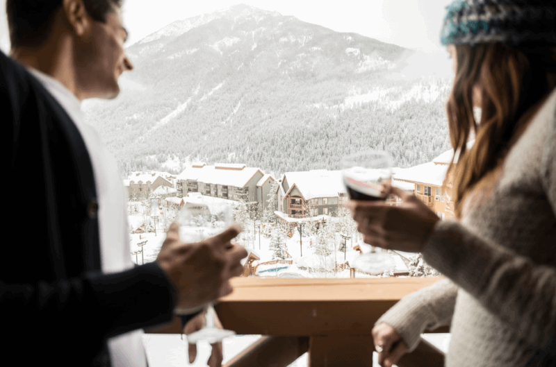 A photo of a man and a woman, each holding a glass of wine, standing on a balcony overlooking Panorama Mountain Resort.