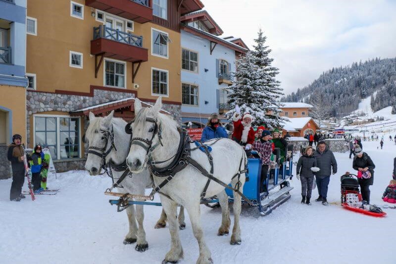 A photo of a team of horses pulling a wagon carrying Santa and children in Sun Peaks Village at Christmas.