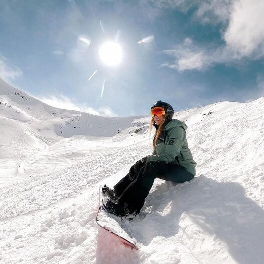 Girl sitting on mountainside with her snowboard and the sun shining behind her